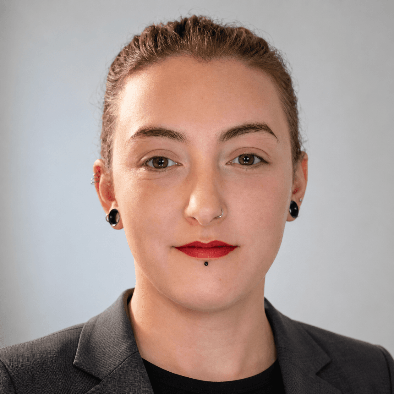 Person with brown hair, red lipstick, nose ring, and black ear gauges wearing a blazer.