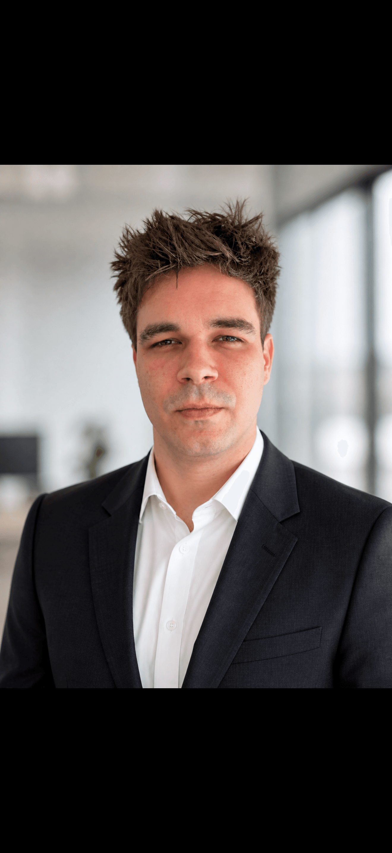 Man with spiky brown hair wearing a dark suit and white shirt in an office.